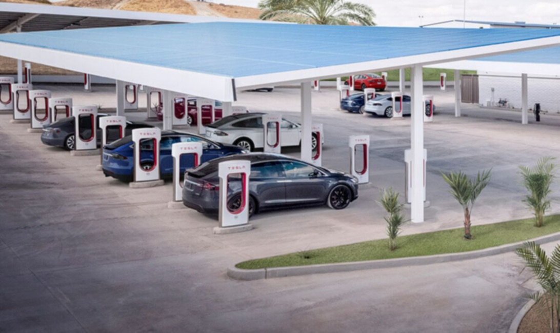 En Tesla Supercharger station med flera laddplatser under ett stort blått soltak, där flera Tesla-bilar laddar.